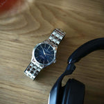 Baume & Mercier Clifton Baumatic 10468 -kello metallirannekkeella, tummalla taululla ja langattomalla kuulokkeella.