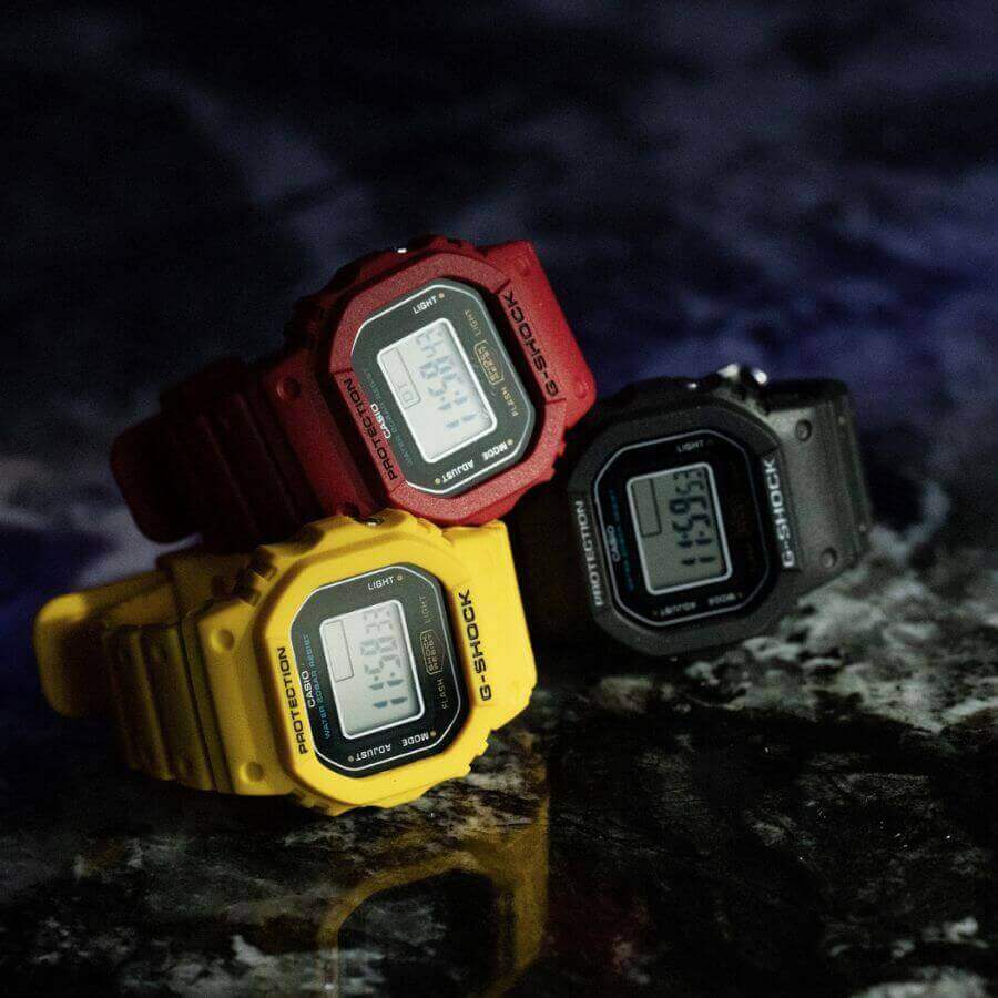 Kolme Casio G-Shock Nano Sormuskello DWN-5600-9ER -kelloa: keltainen, punainen ja musta, taustalla tummat pinnat.