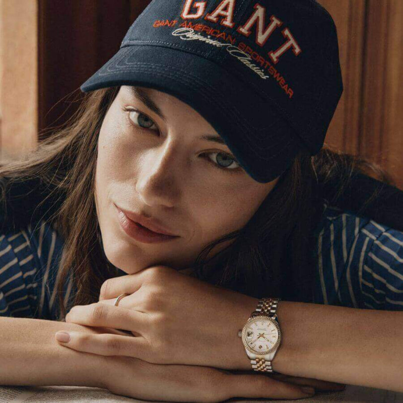 Naisen kädessä Gant Sussex Mini Bijou Rannekello G181103, musta lippalakki päässä, hymyilevä ilme.